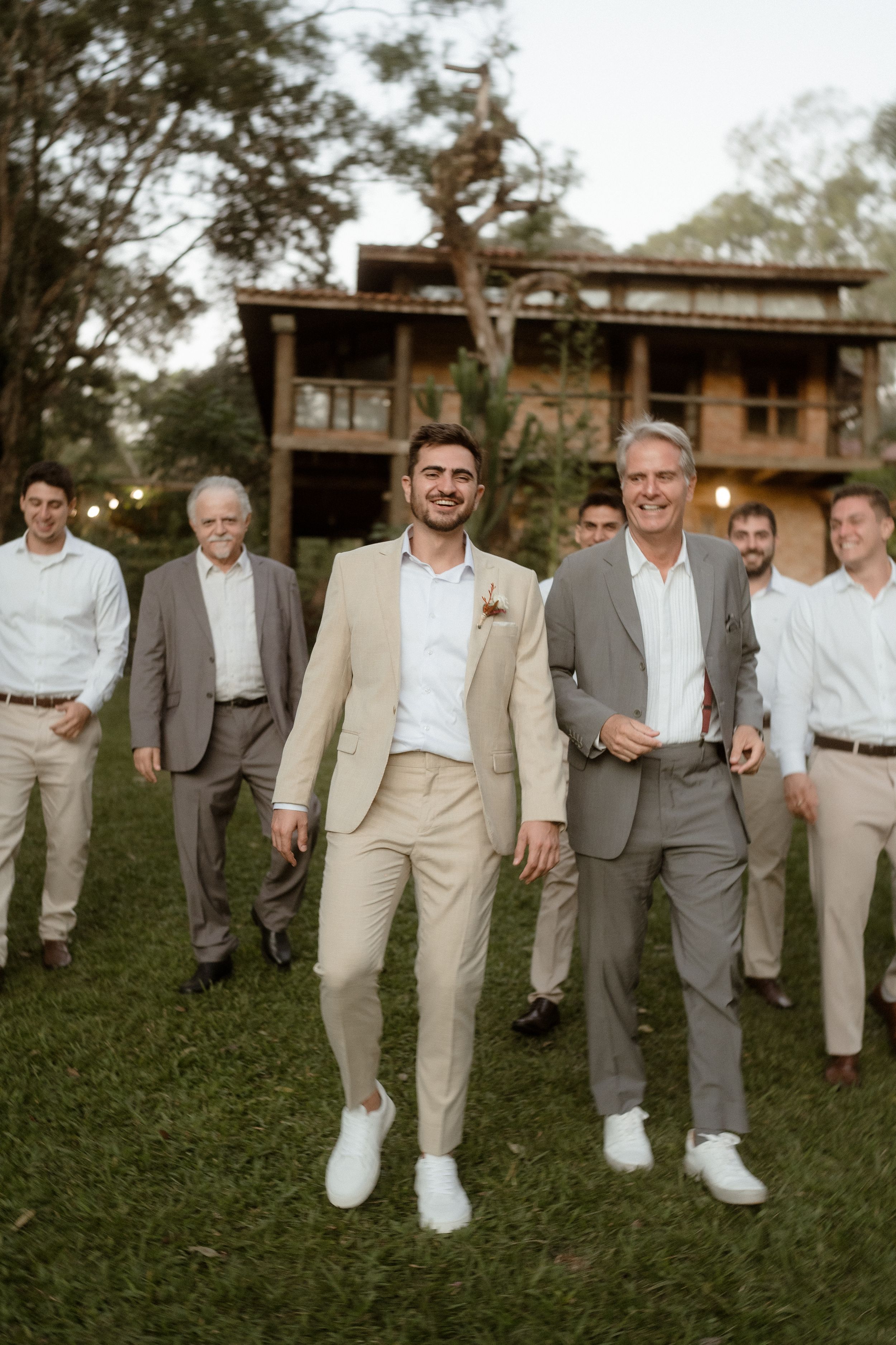 Wedding party in light suits poses casually on grass in front of rustic wooden building.