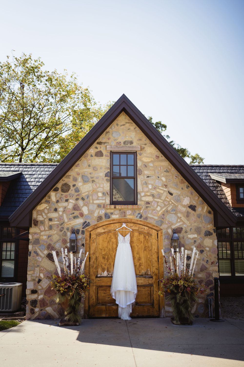 White wedding dress hanging on wooden door of stone chapel with flanking floral arrangements.