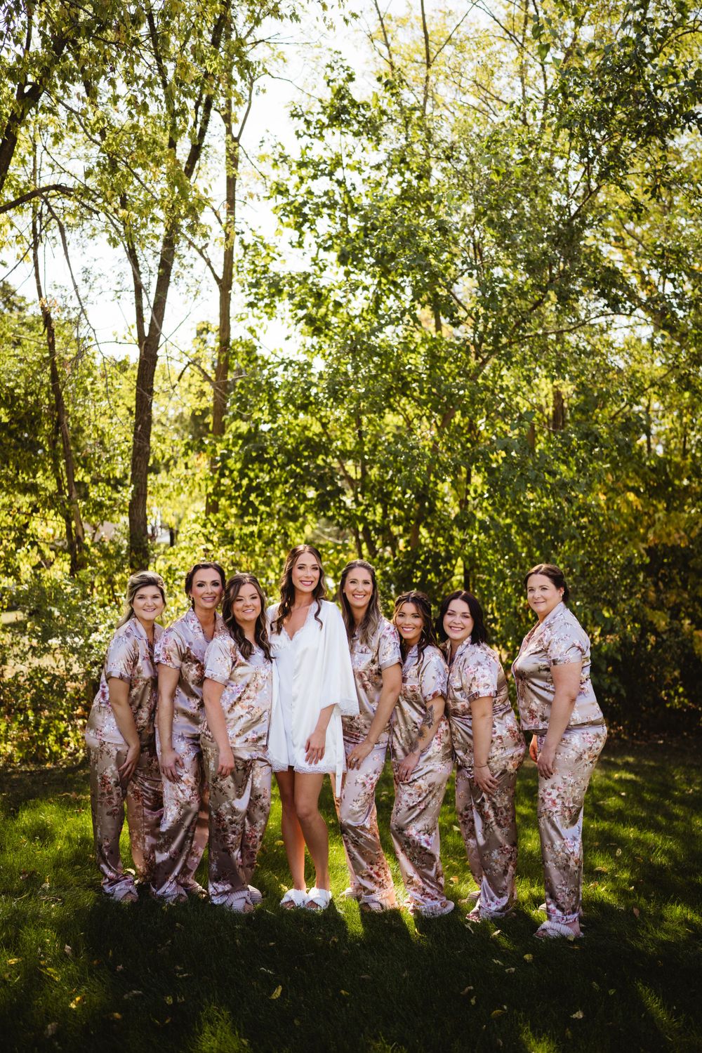 Bride in white robe surrounded by bridesmaids wearing matching floral robes in a sunny garden setting.
