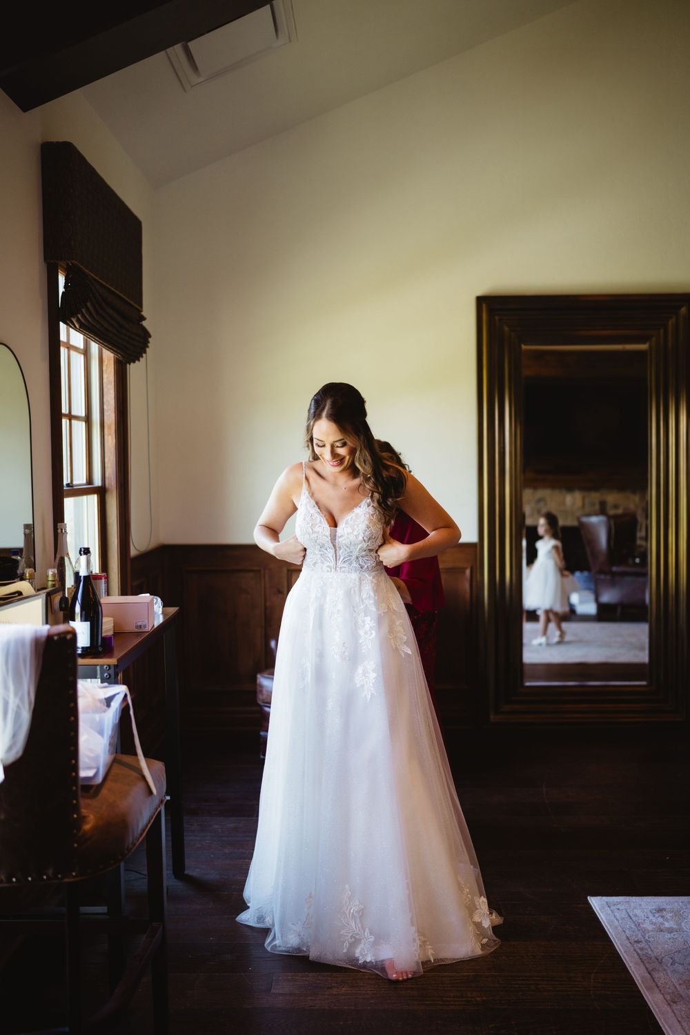 Bride in elegant lace wedding gown stands in vintage room with dark wood floors and natural lighting.