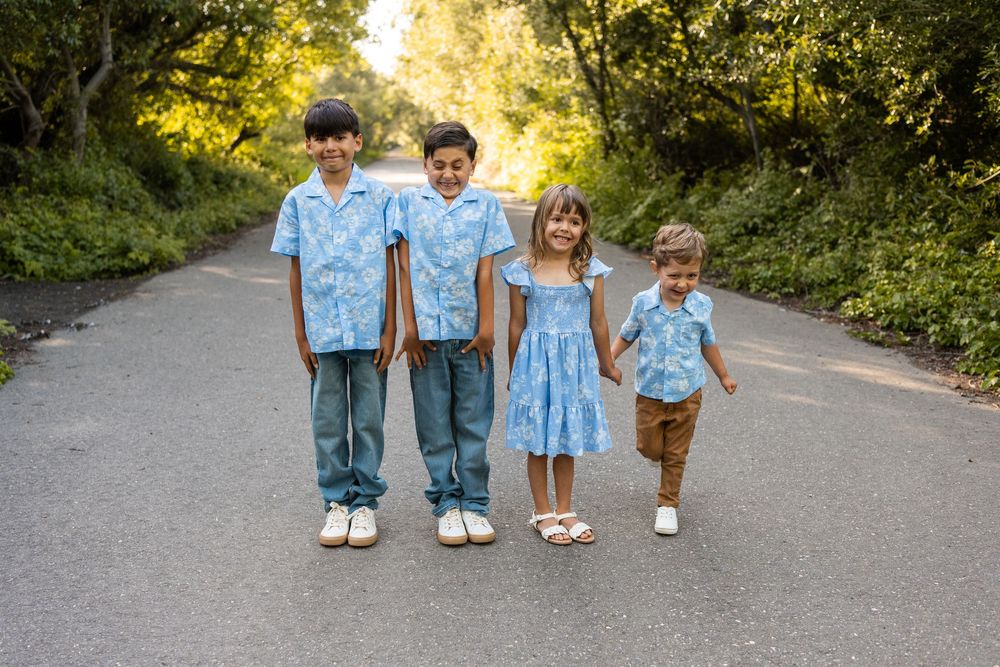 Four children in matching blue floral outfits pose together on a tree-lined road for a family photo.