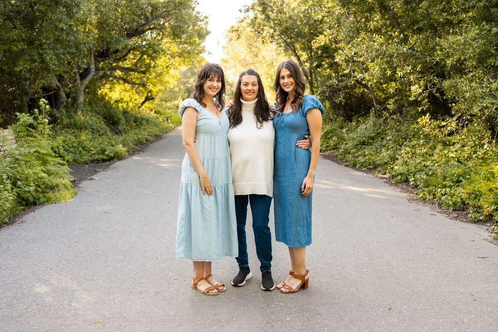 Three smiling women in blue dresses and white sweater pose together on a tree-lined path outdoors.