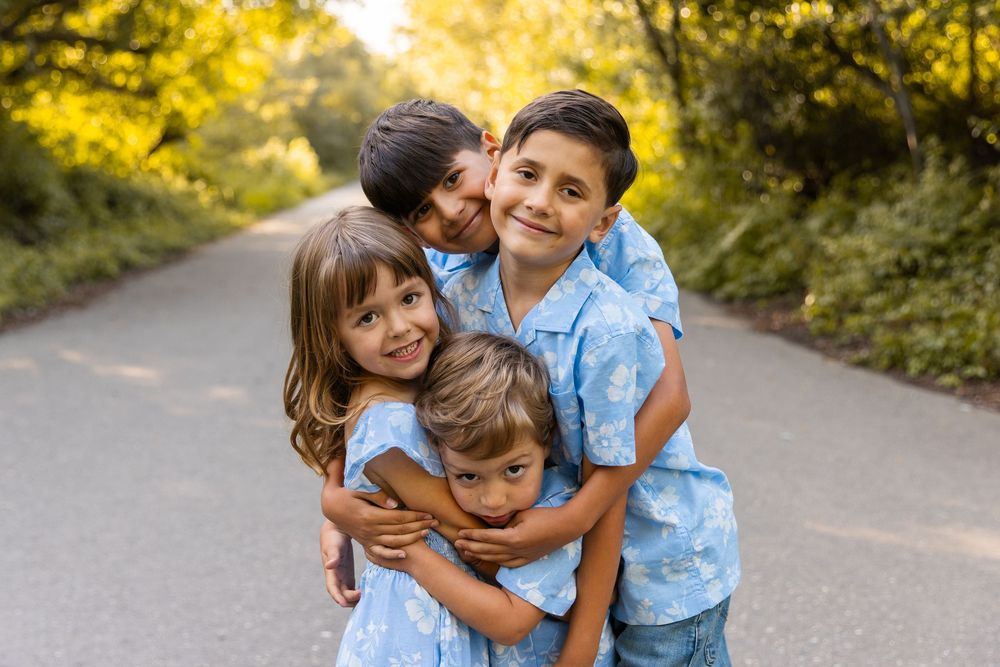 Four young siblings in matching blue outfits hugging and smiling on a tree-lined path during a family photo session.