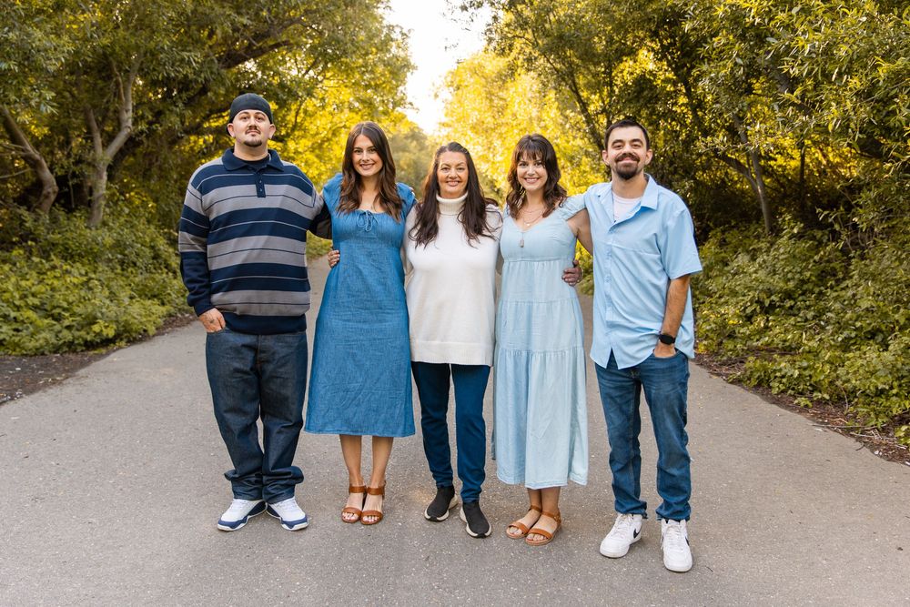 Five smiling adults pose together on a tree-lined path during golden hour in a casual outdoor family portrait.