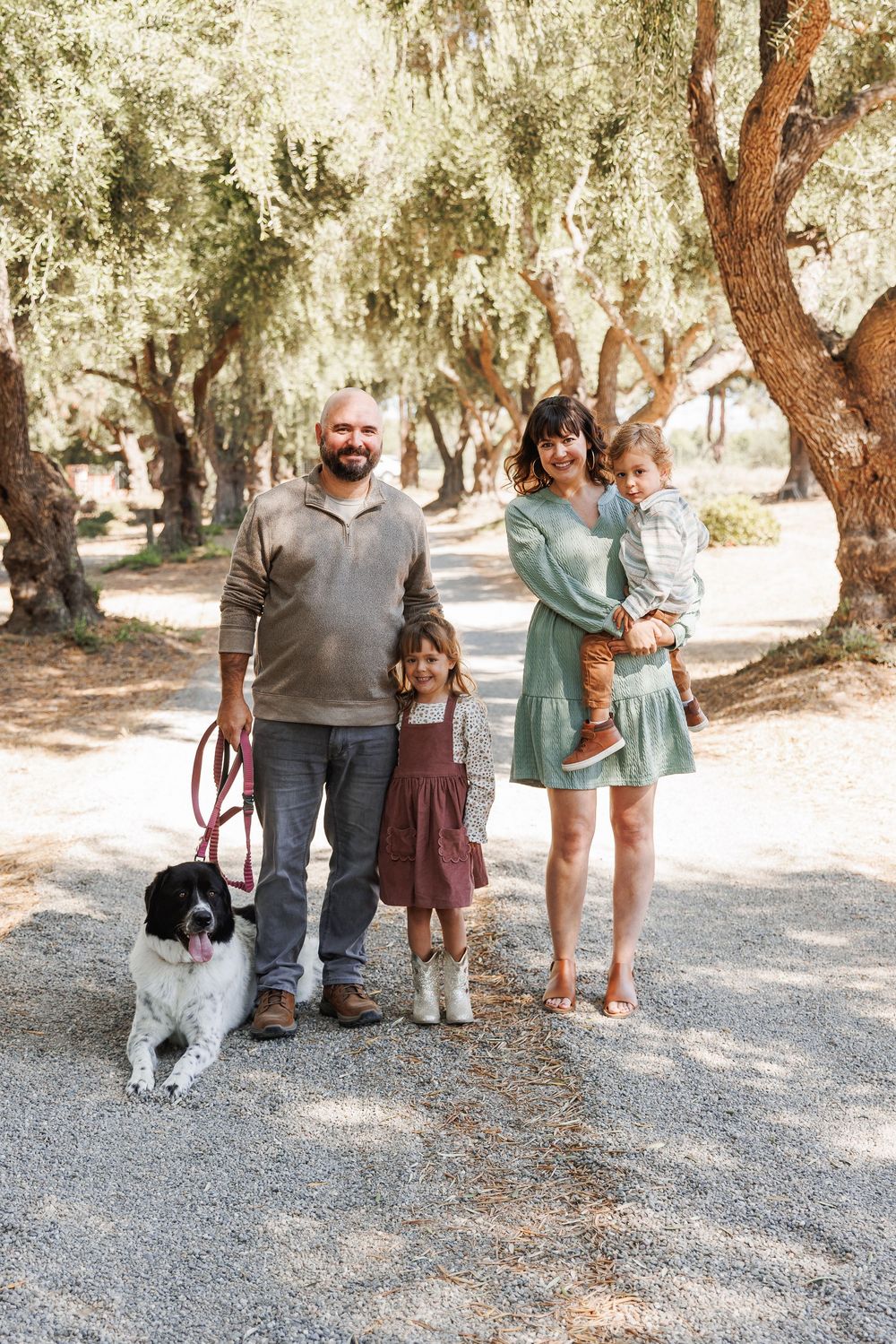 Family portrait with two children and a dog standing on a gravel path under olive trees.