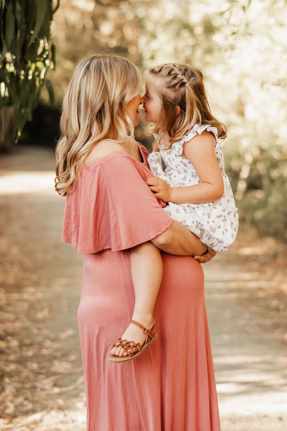 Pregnant blonde mother in pink dress touching noses with young daughter outdoors.