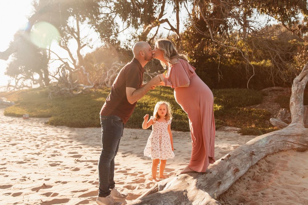 Couple kissing at golden hour beach while young daughter watches in foreground.