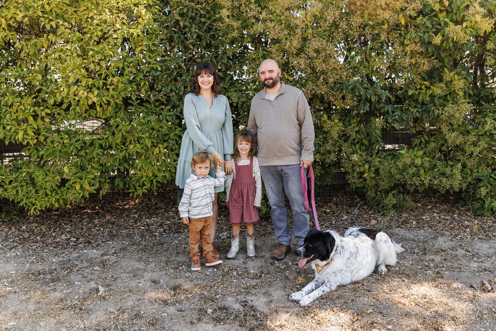 Family of four with a dog posing outdoors in front of lush green hedges on a sunny day.