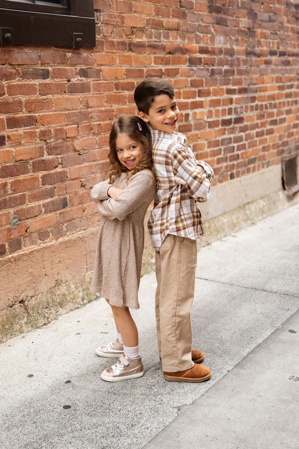 Two children standing back-to-back with arms crossed, smiling in front of a rustic brick wall.