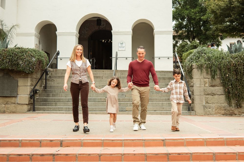 Family of four holding hands on steps outside a white building.