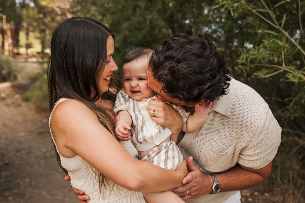 A happy young family with a mother holding her smiling baby while the father kisses the child outdoors.