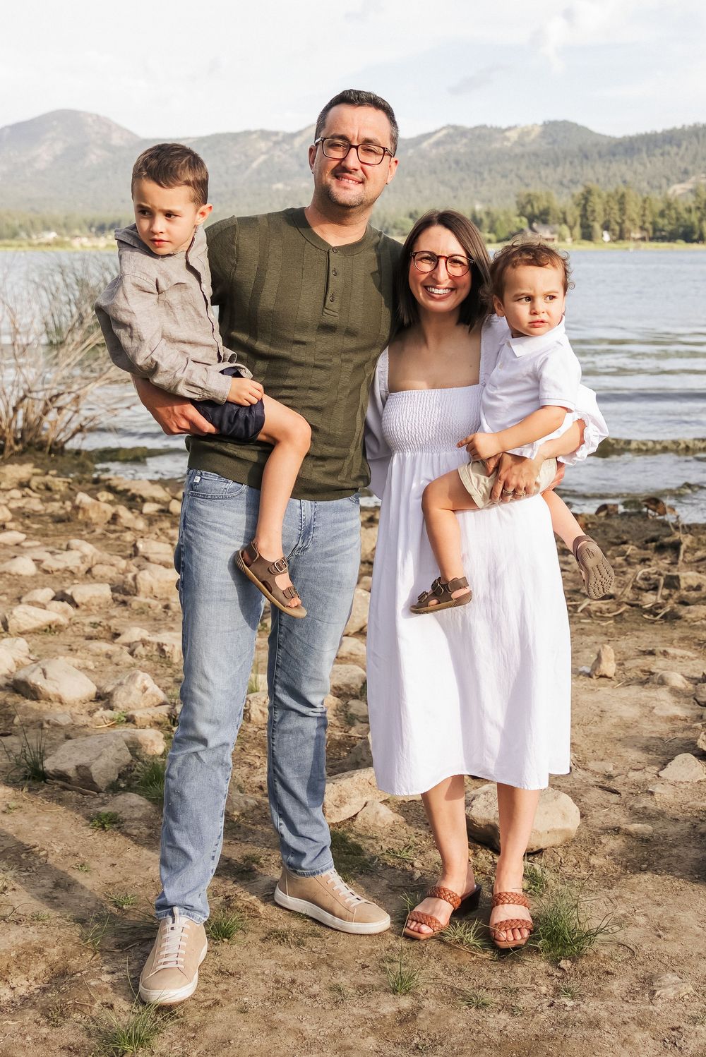 A happy family of four poses by a mountain lake, parents holding two young boys, dressed in casual outdoor attire.