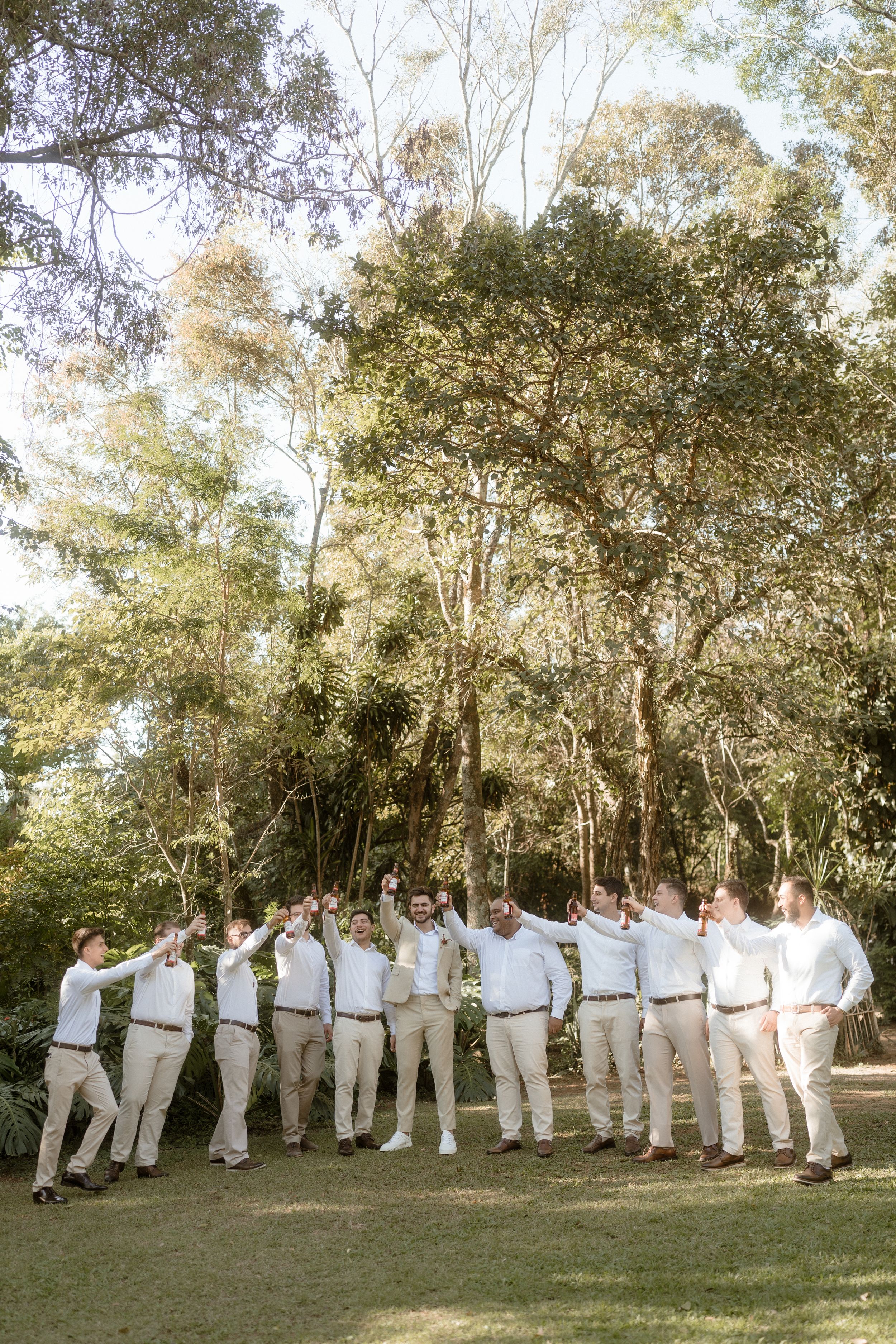 Wedding party in white shirts and khaki pants posing together under tall trees.
