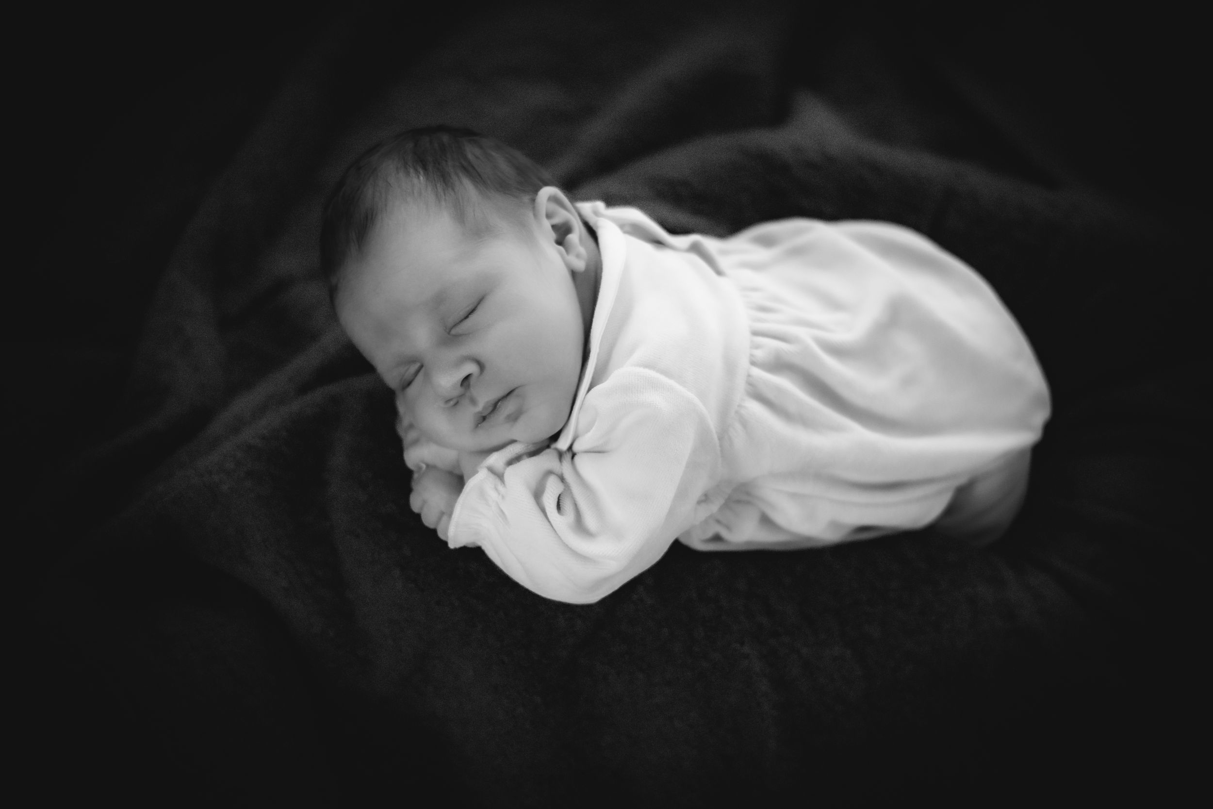 Peaceful sleeping newborn baby in white onesie on dark fabric.