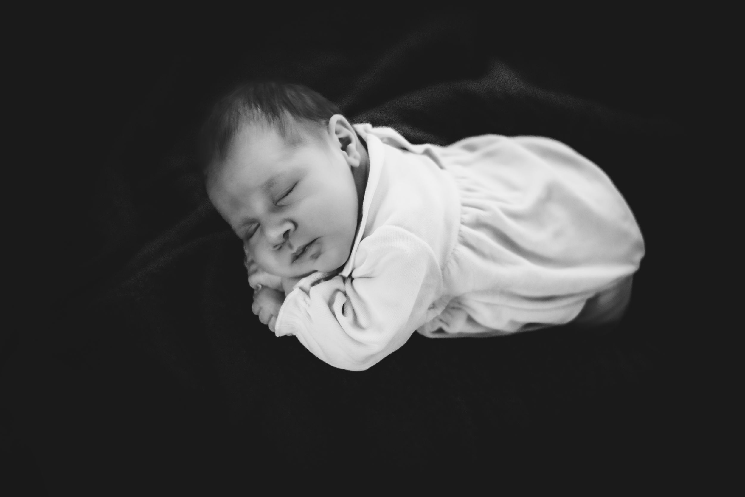 Black and white close-up portrait of a sleeping infant in white clothing against dark background.