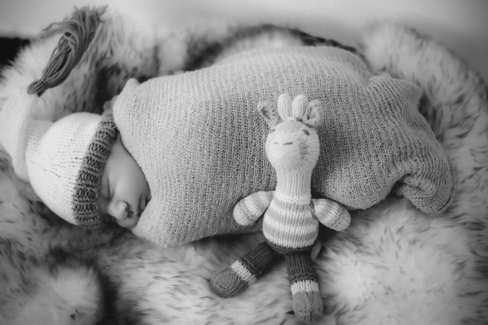 Black and white photo of sleeping baby with knit toy on soft fur blanket.