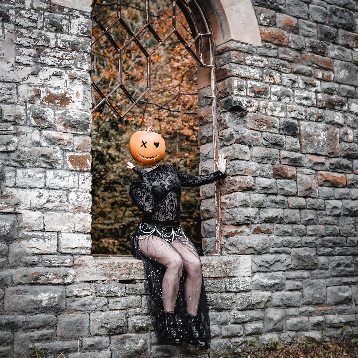 Figure in black lace dress with pumpkin head sits in stone archway during autumn season.