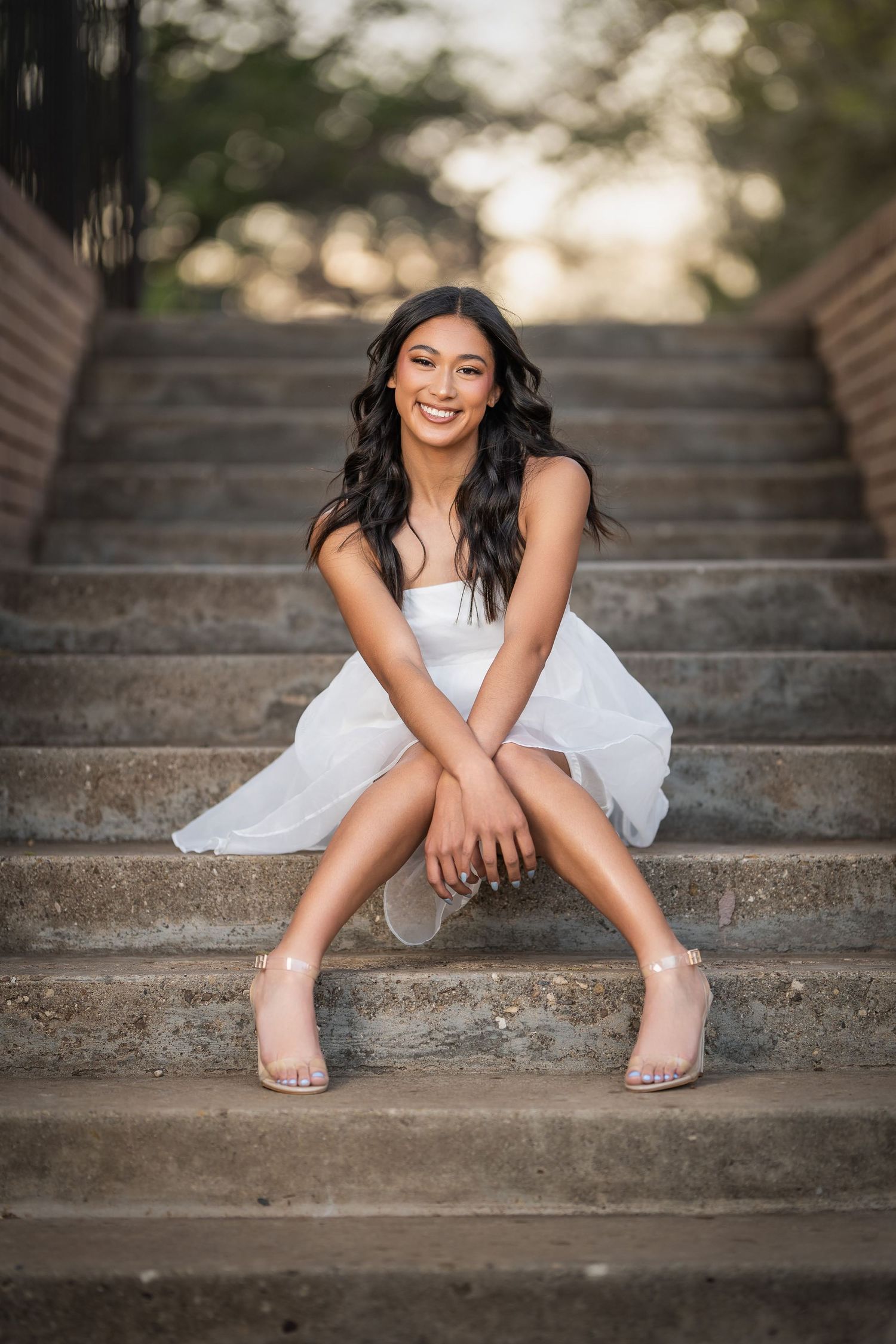 A person in a white dress sits playfully on concrete steps during a portrait photoshoot with natural golden lighting.