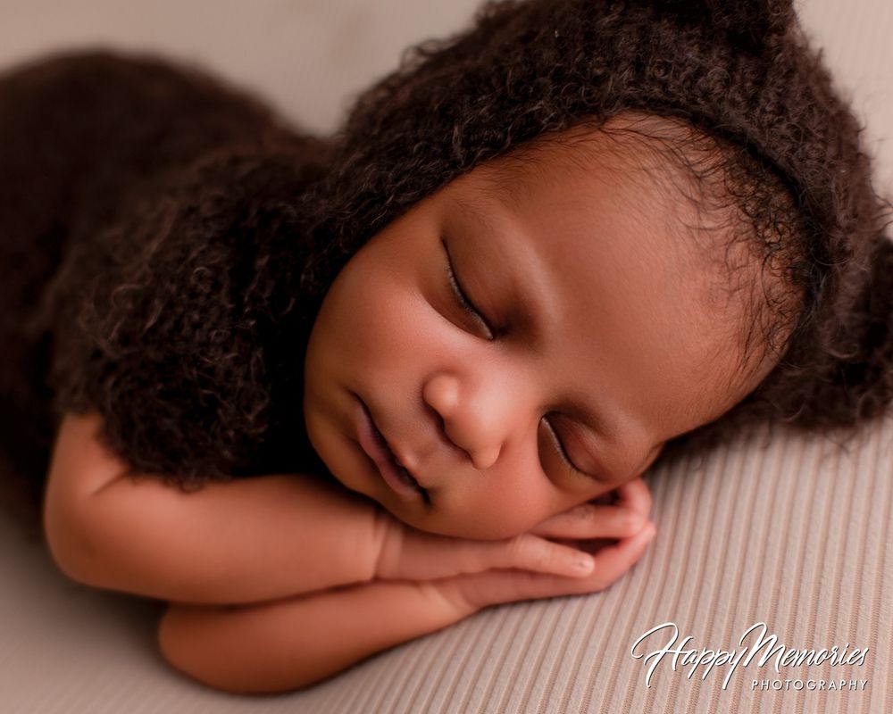 Sleeping newborn baby in a brown knit bear bonnet and wrap, resting its cheek on folded hands during a soft newborn photography session.
