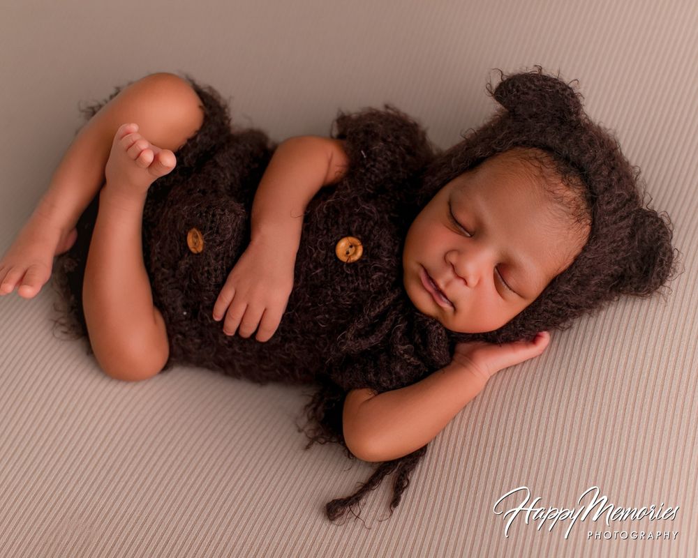 Sleeping newborn baby in a brown knit bear bonnet and matching romper with wooden buttons, posed on a soft beige blanket.