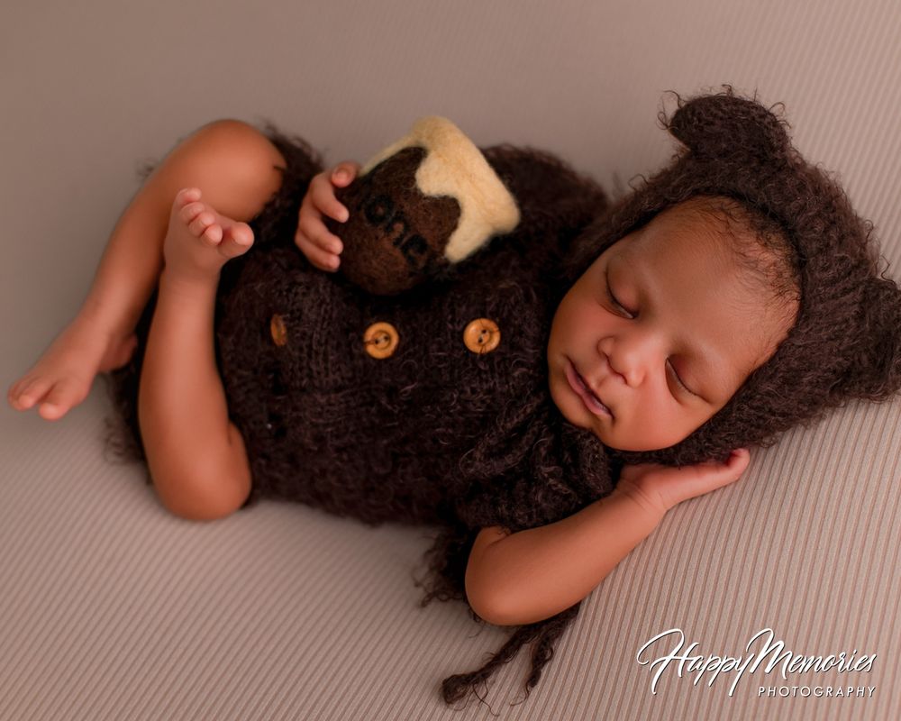 Sleeping newborn baby in a brown knit bear bonnet and outfit with wooden buttons, resting on a soft gray backdrop beside a teddy bear.