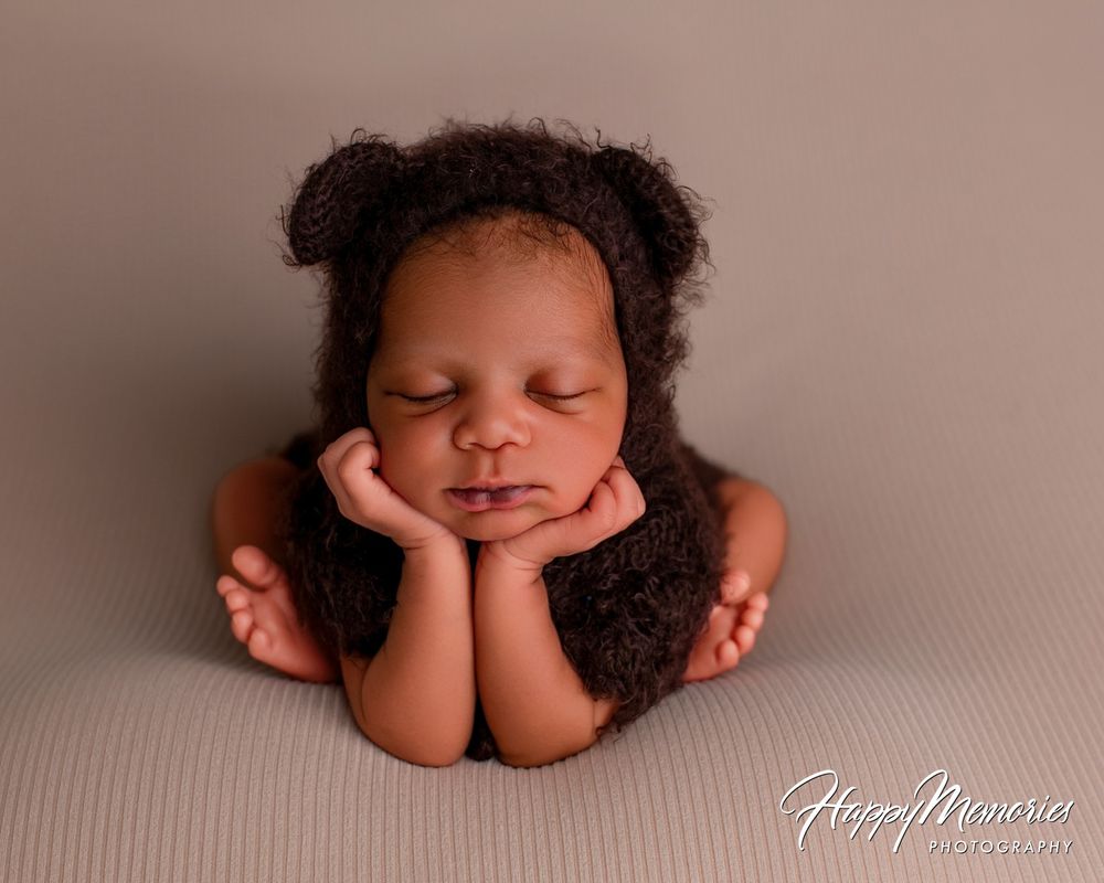 Newborn baby in a fuzzy brown bear-ear bonnet sleeping peacefully in a froggy pose with chin resting on hands on a neutral backdrop.