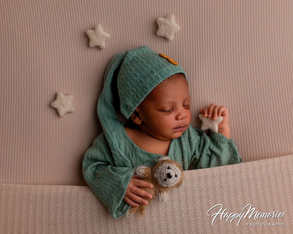 Sleeping newborn baby in a green knit nightcap and outfit, holding a tiny teddy bear and star, tucked under a blanket in a newborn photo.