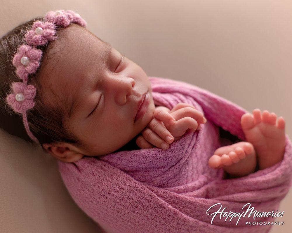 Sleeping newborn baby girl wrapped in a pink knit blanket wearing a pink flower headband, captured in a newborn photography session.