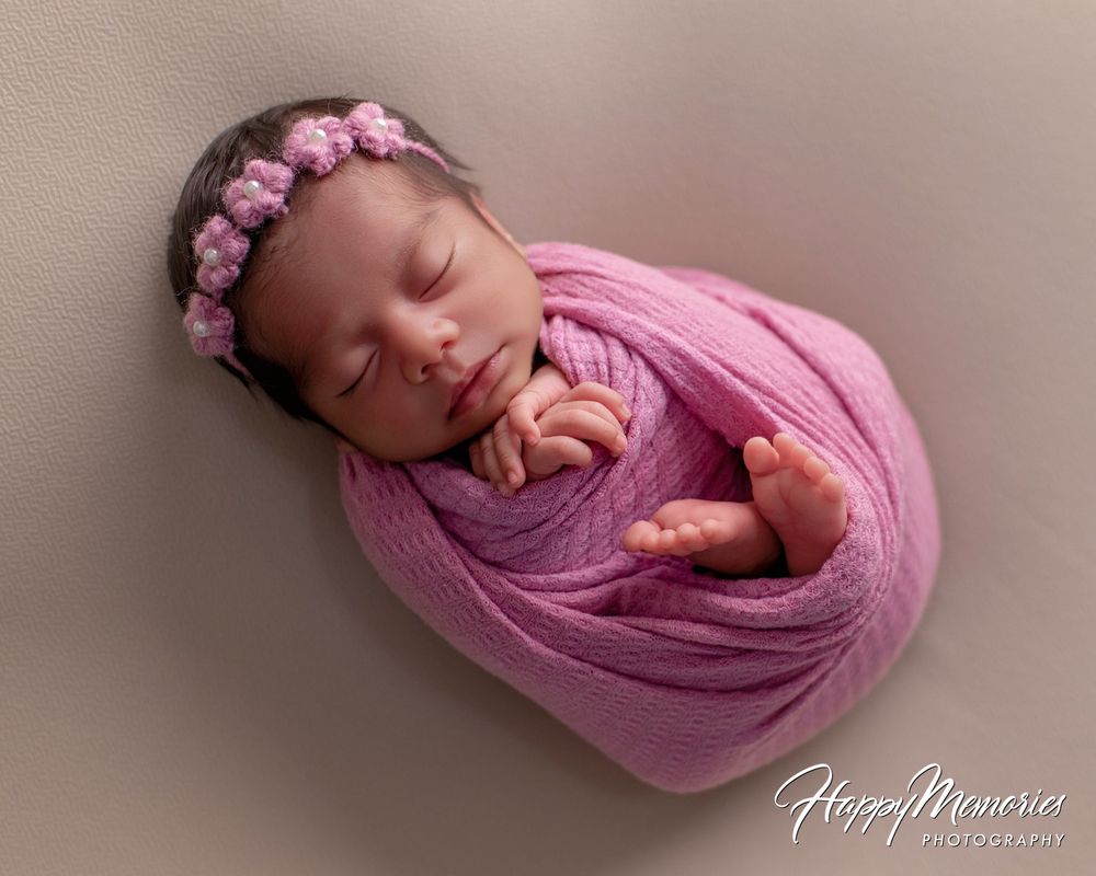 Sleeping newborn baby girl wrapped in a pink knit swaddle wearing a floral headband in a soft newborn photography portrait.