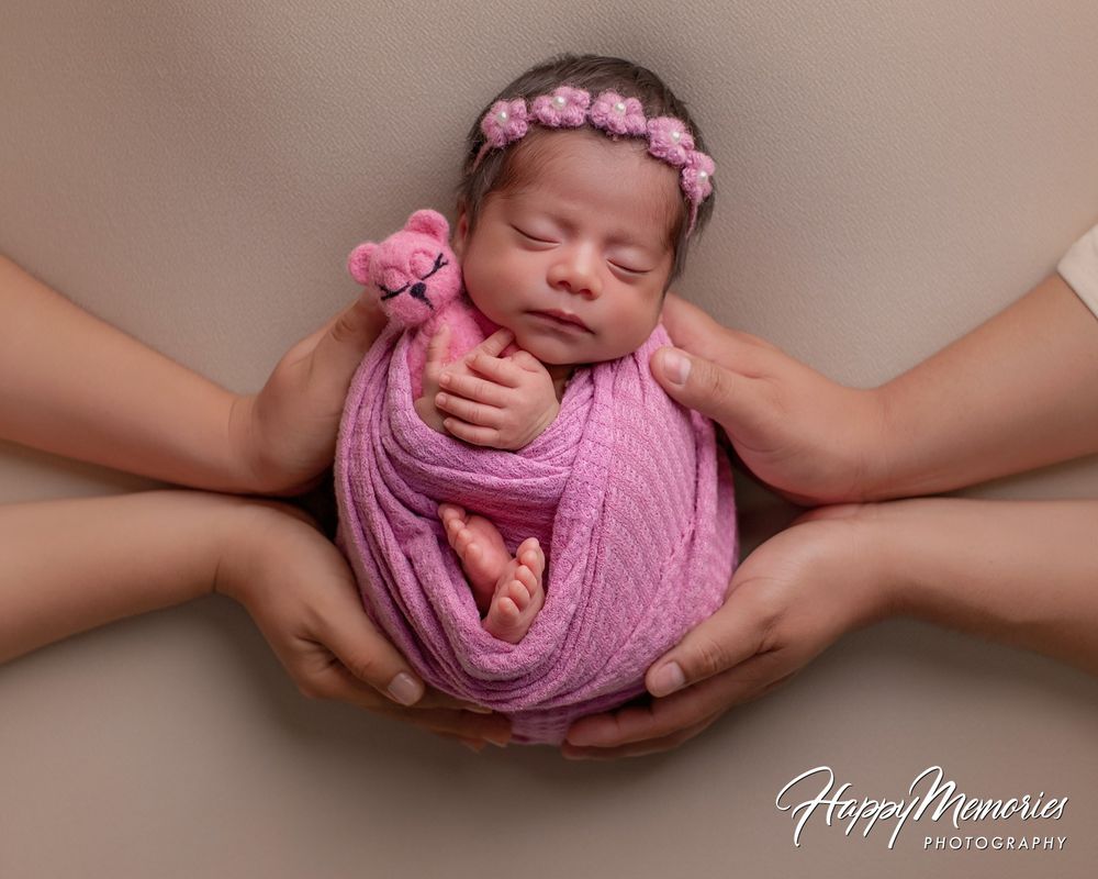 Sleeping newborn baby wrapped in pink swaddle with floral headband holding a tiny pink teddy bear, cradled in four parents' hands.