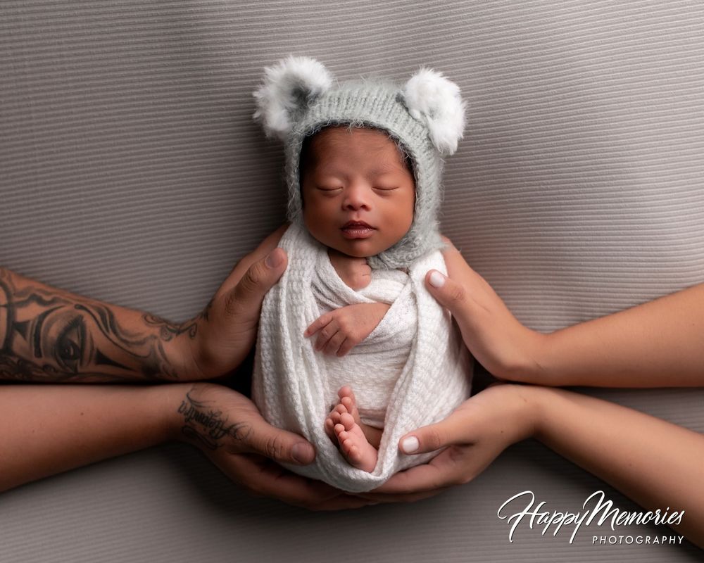 Newborn baby in a gray knit bear-ear hat sleeping while cradled by four parent hands, a Happy Memories Photography newborn portrait.