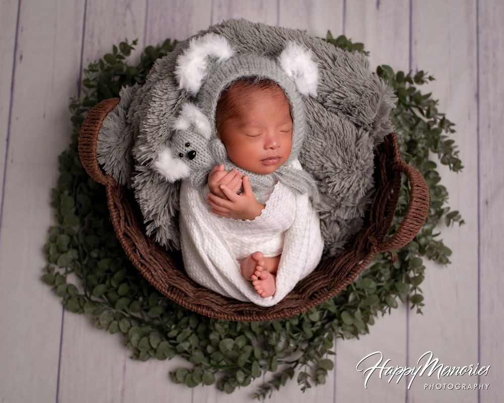 Sleeping newborn baby in a koala bear hat swaddled in white wrap, resting in a wicker basket with greenery on a rustic wood floor.