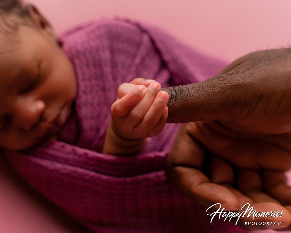 Newborn baby wrapped in a purple knit blanket gently holding an adult's finger against a soft pink background.