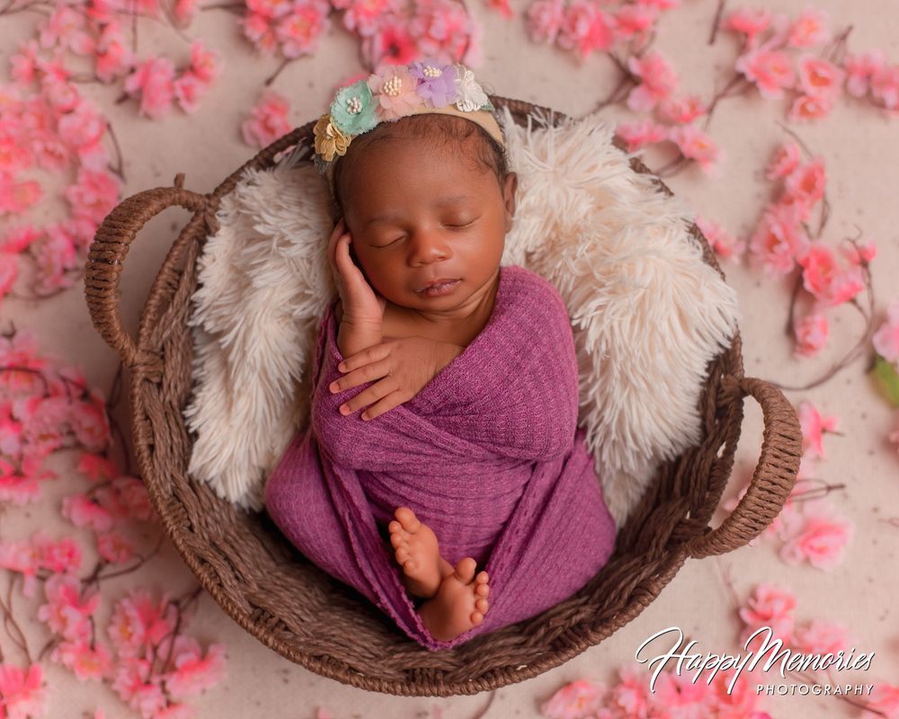 Sleeping newborn wrapped in purple swaddle with a floral headband resting in a wicker basket on a pink petal-covered backdrop.