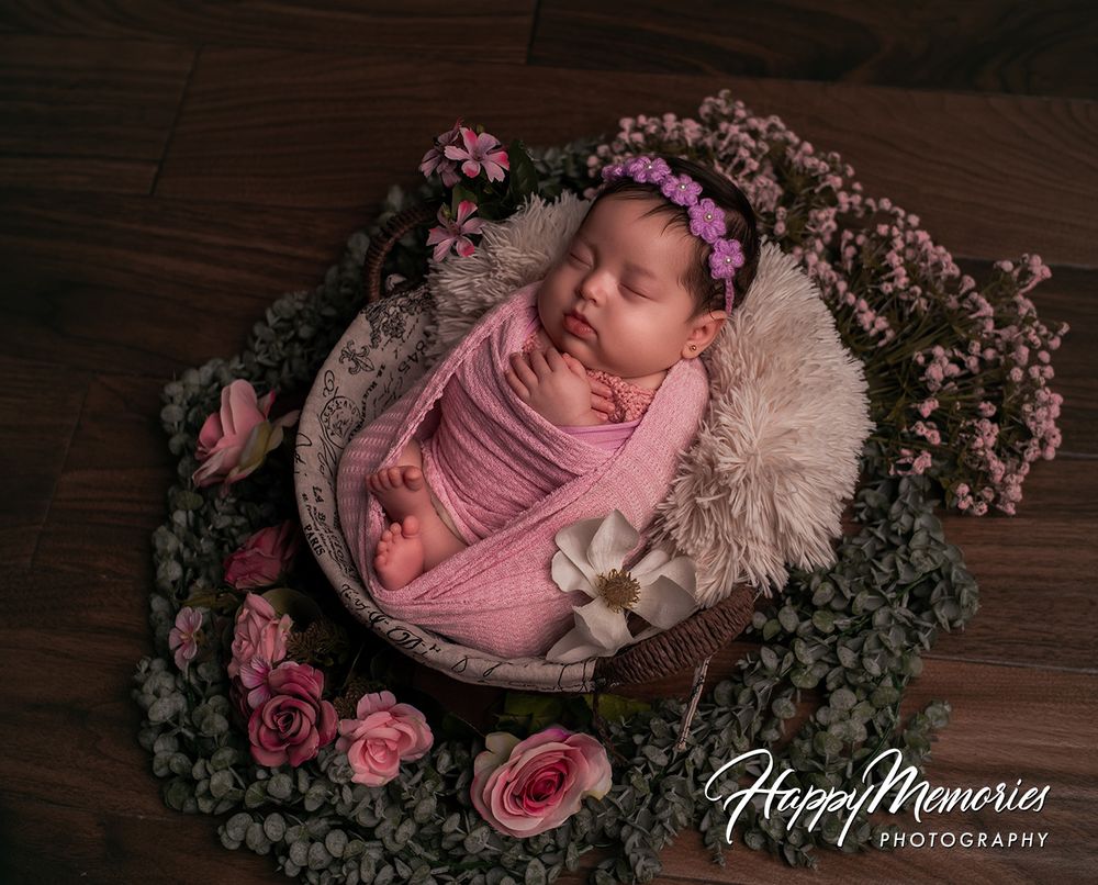 Peaceful newborn girl wrapped in pink swaddle with floral headband sleeping in wooden bowl surrounded by roses.