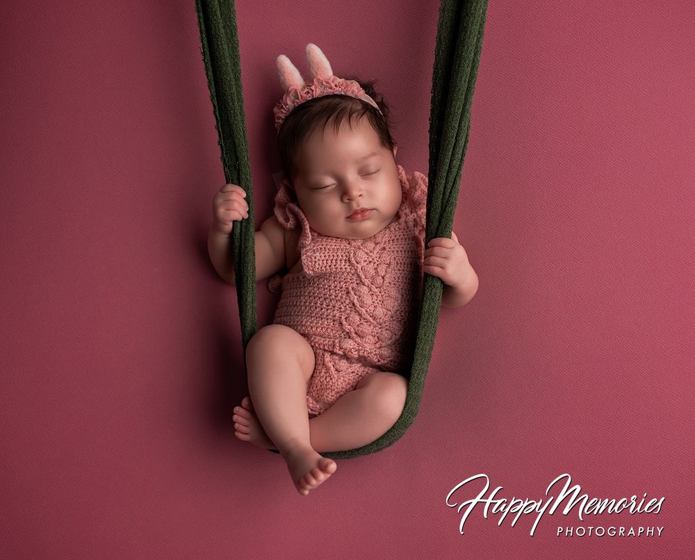 Newborn baby in pink knit romper and bunny headband sleeping peacefully in a green fabric swing on pink background.