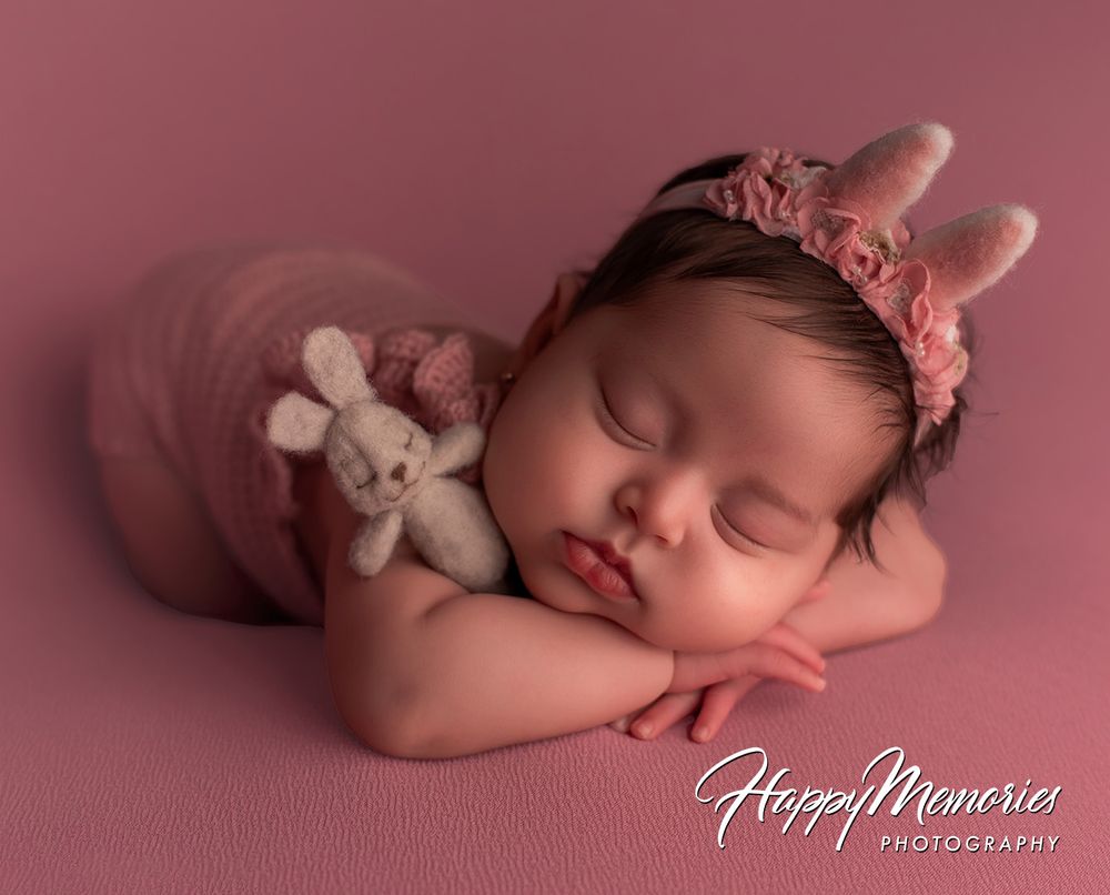 Sleeping newborn baby girl in pink outfit with bunny headband hugging a small knit bunny toy on pink backdrop.