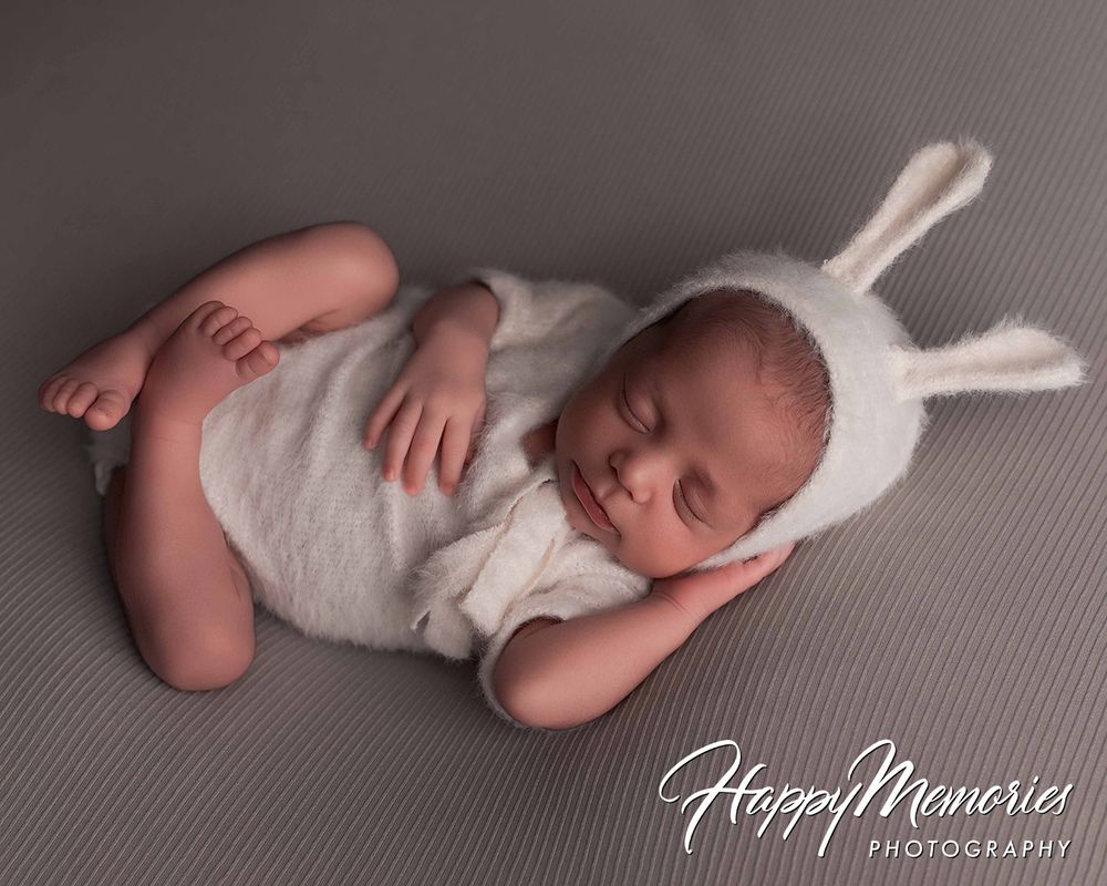 Sleeping newborn baby in white bunny romper and hat on gray backdrop, Happy Memories Photography.