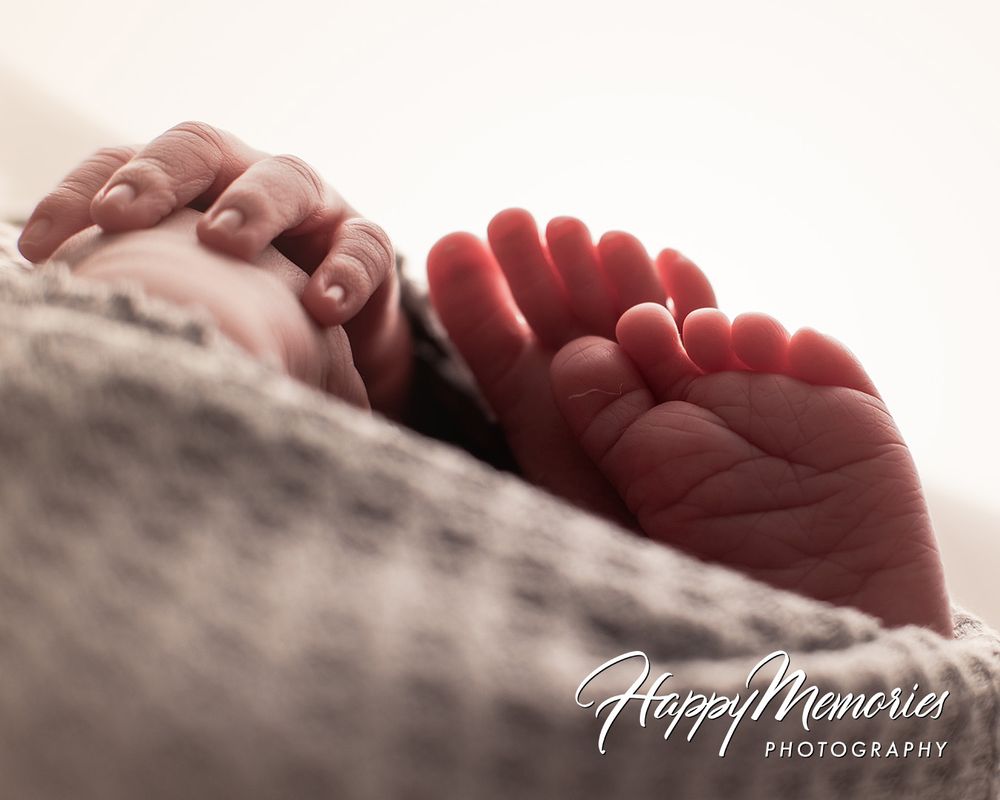 Close-up of a newborn baby's tiny hands and feet wrapped in a soft gray blanket, captured by Happy Memories Photography.