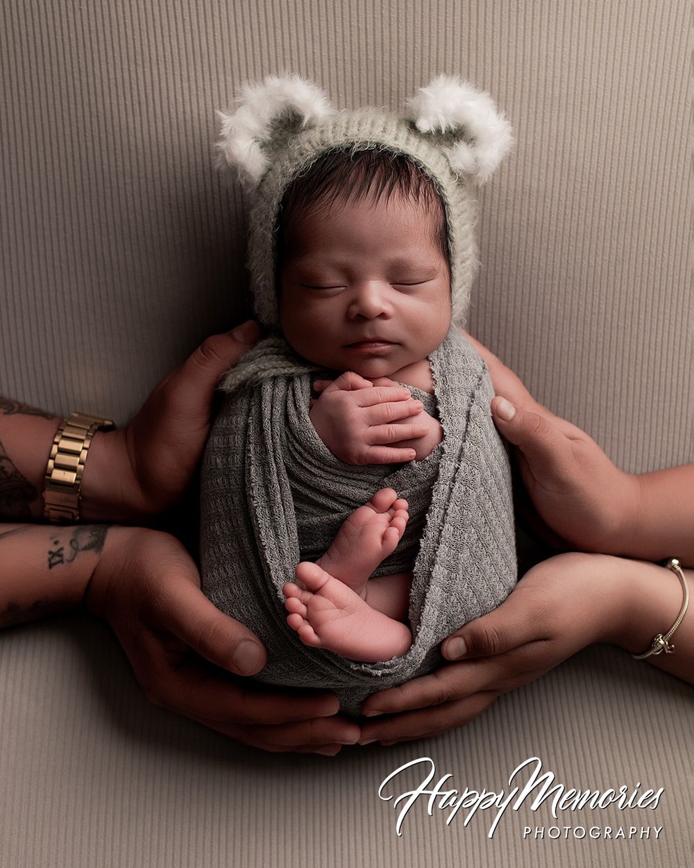 Newborn baby wrapped in gray swaddle with bear ear bonnet, cradled in parents hands, professional newborn photography.