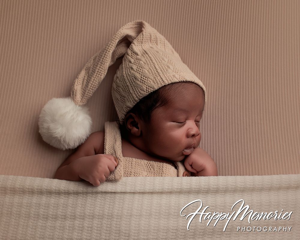 Sleeping newborn baby wearing a beige knit hat with pompom and matching romper on a tan background.