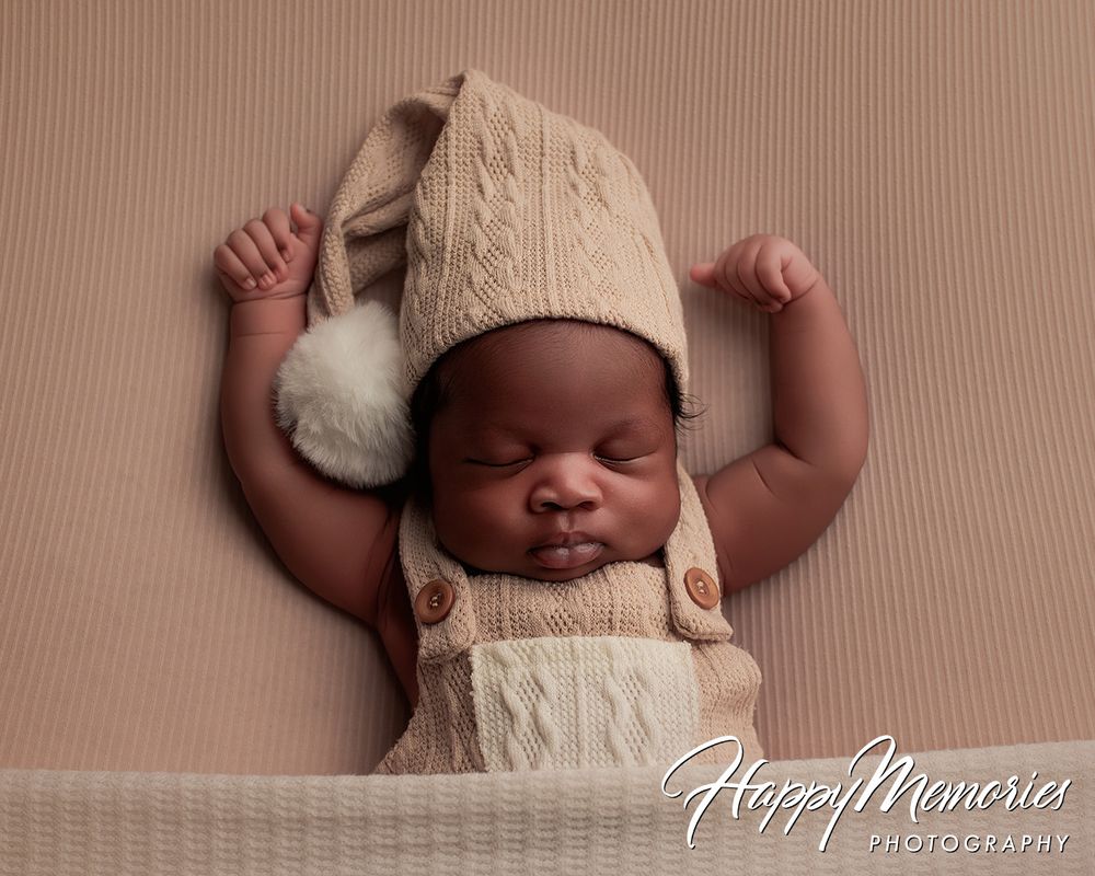 Newborn baby in beige knit hat and romper stretching arms wide while sleeping peacefully on tan backdrop.