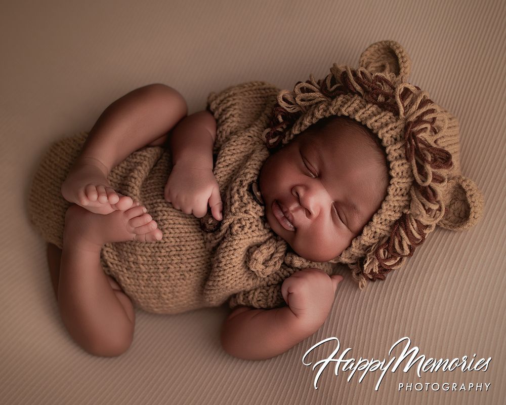 Smiling newborn baby in a brown knit lion costume with mane bonnet, posed on a tan backdrop.