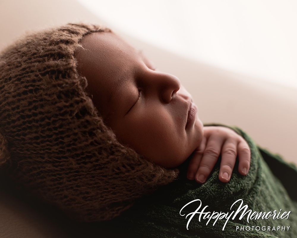 Peaceful newborn baby sleeping in a brown knit bonnet and green wrap, captured by Happy Memories Photography.