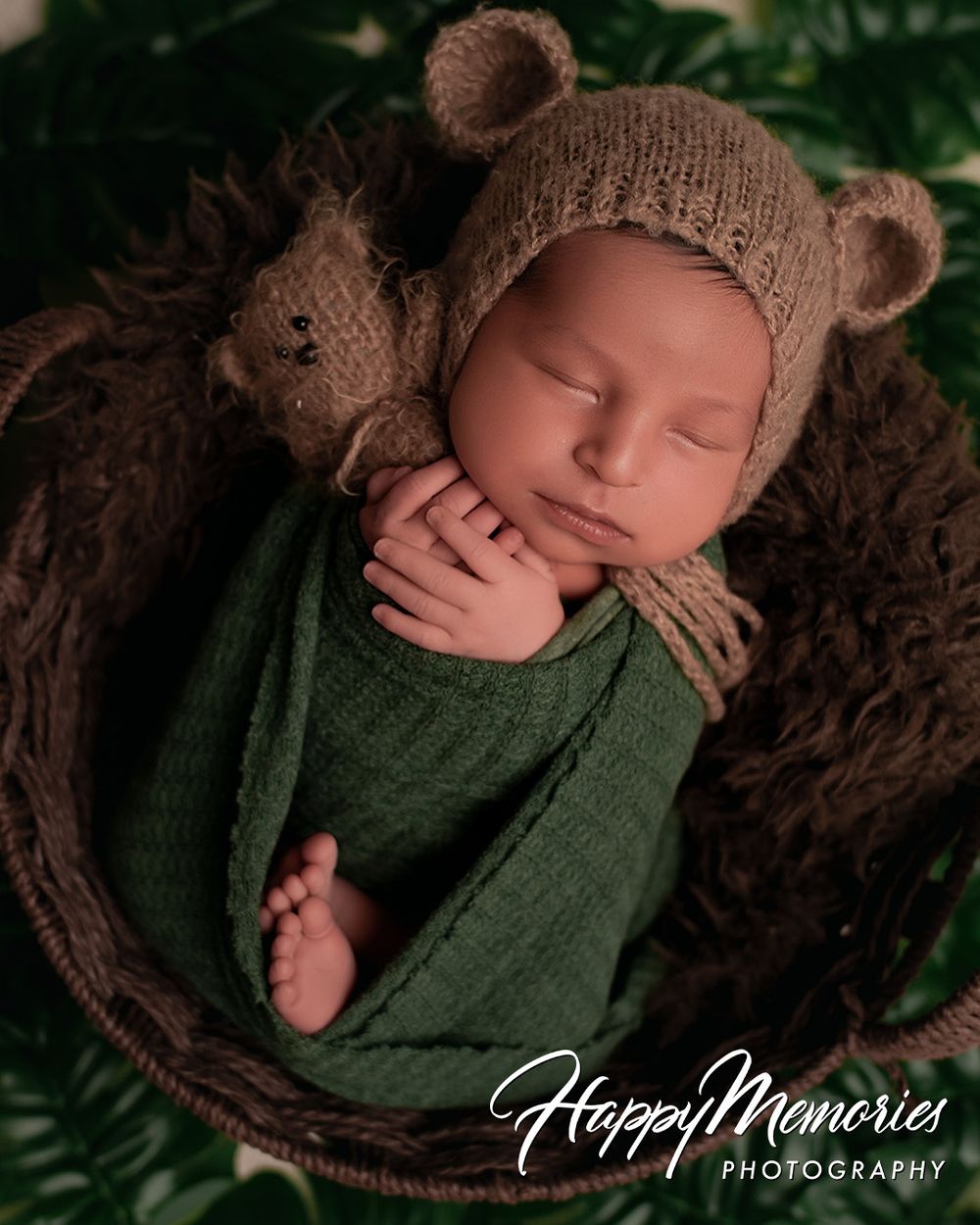 Sleeping newborn baby wrapped in green swaddle wearing knit bear bonnet nestled in wicker basket with teddy bear.