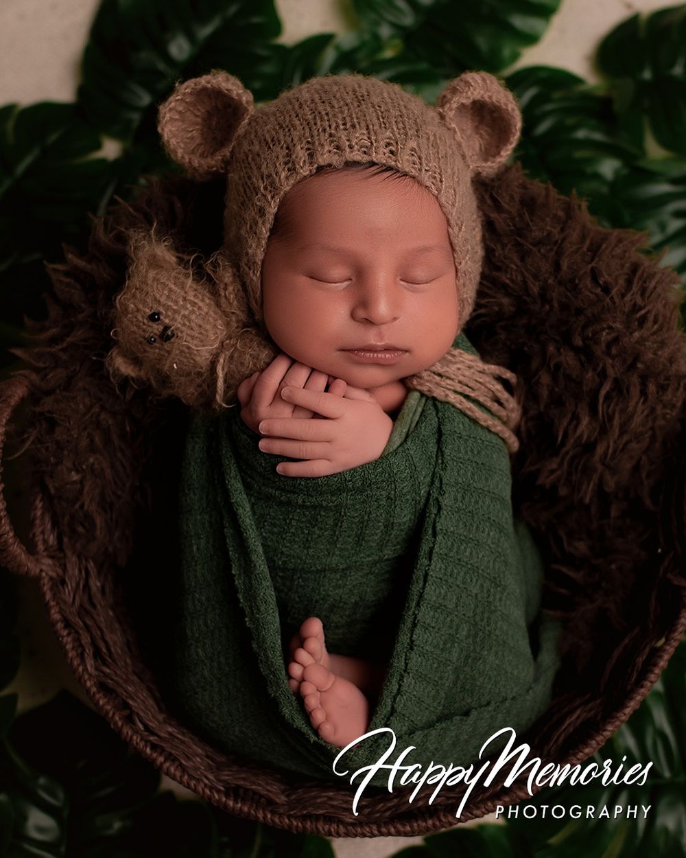 Newborn baby wrapped in green swaddle wearing knit bear bonnet, sleeping peacefully in wicker basket with stuffed bear.