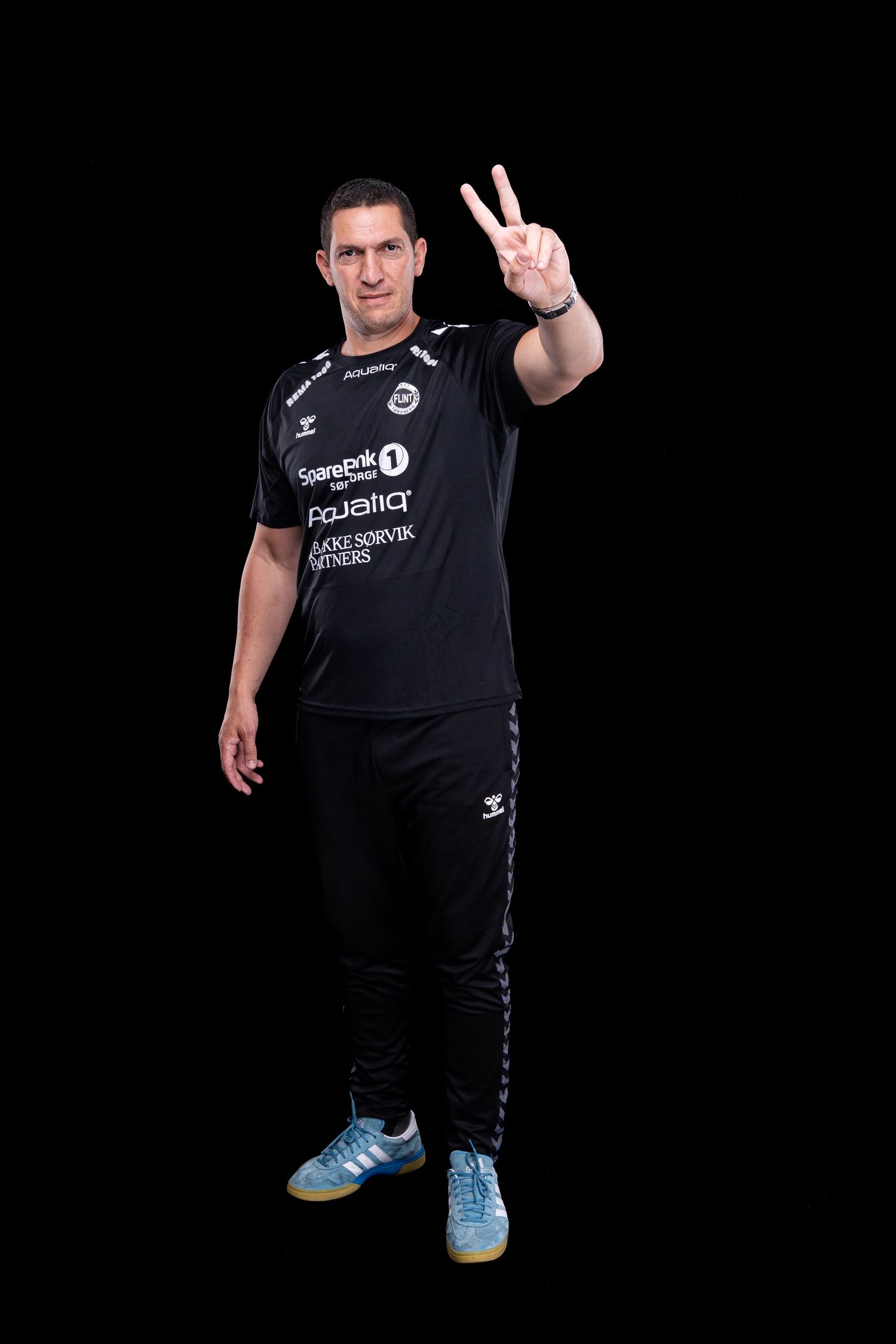 Athletic person in black sportswear and blue shoes poses against dark background making peace sign gesture.