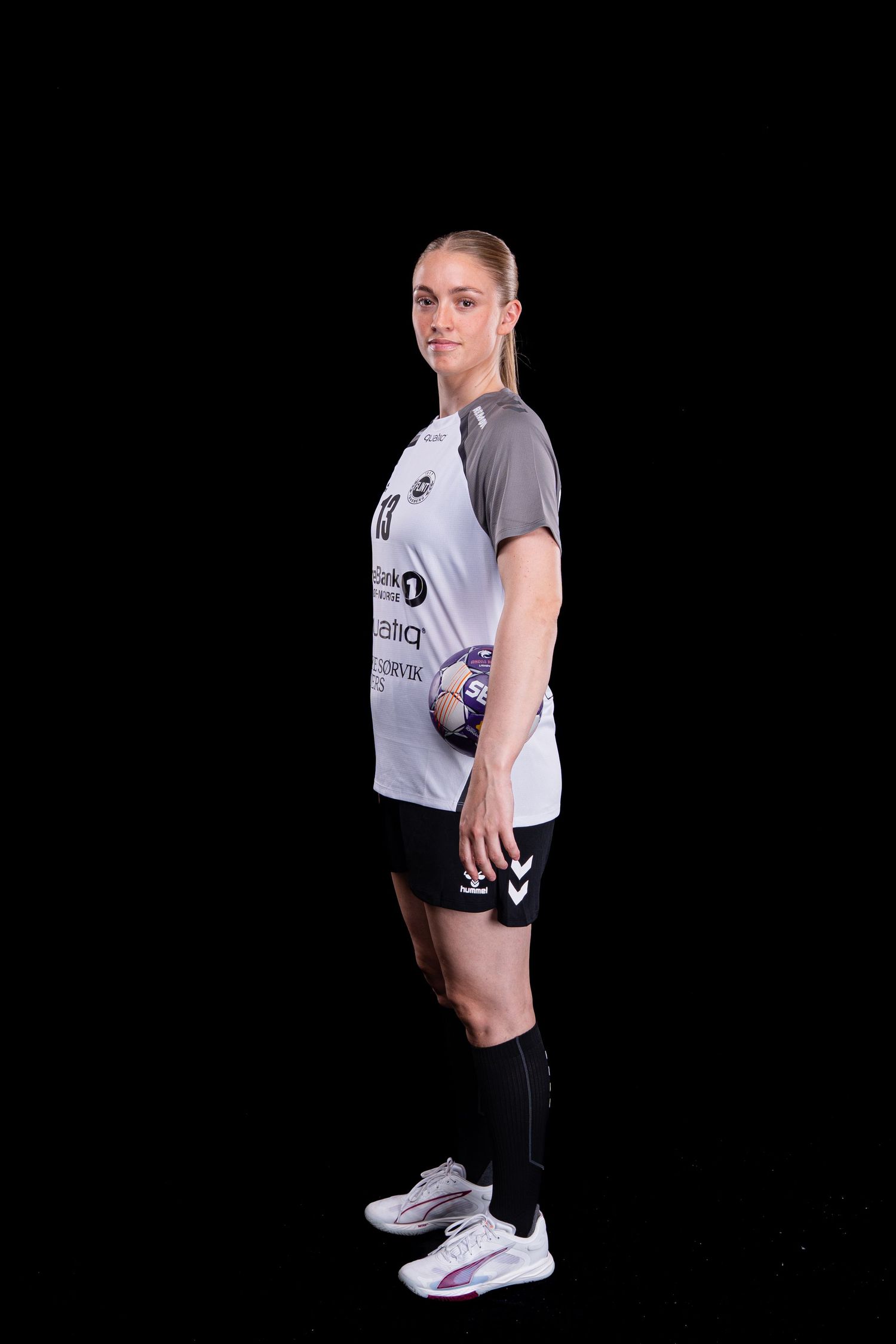 Athletic figure in grey t-shirt and black shorts poses in professional stance against dark backdrop wearing white sneakers.