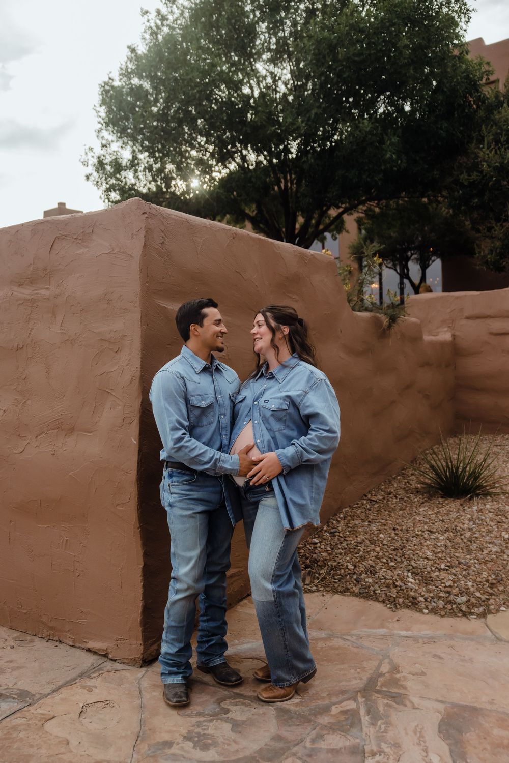 Happy expecting couple in matching denim standing against adobe wall, hands on pregnant belly, smiling at each other.