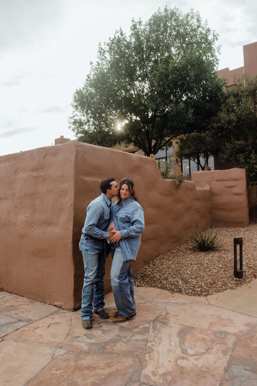 Expecting couple in coordinating denim outfits embracing near rustic adobe wall with lush green tree backdrop.